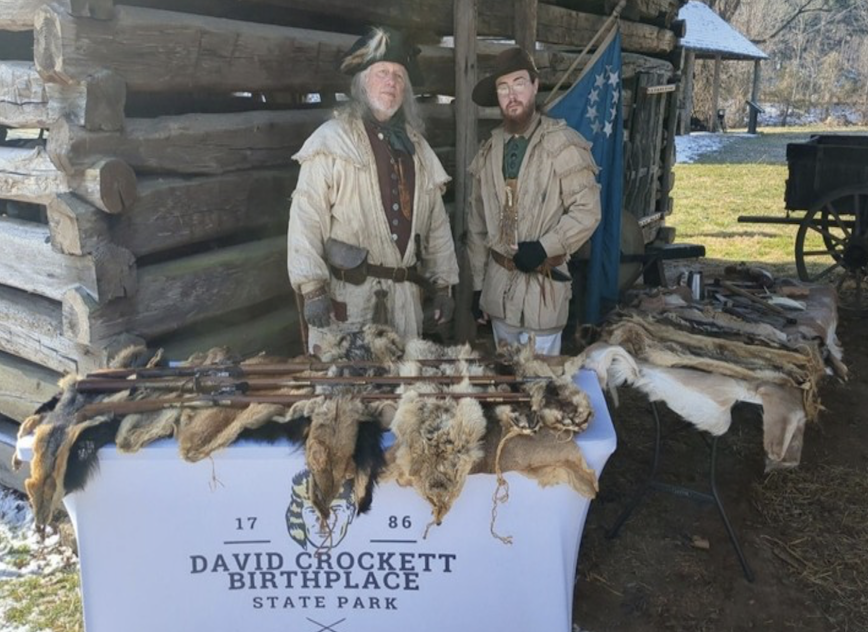 Two frontier reenactors stand beside a log cabin with a display of pelts and rifles at David Crockett Birthplace State Park.